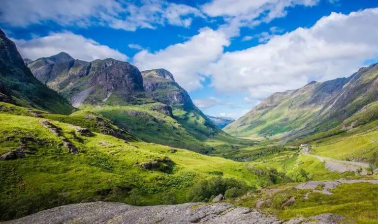 Highland valley with steep mountains, winding road and lush green slopes under blue sky with white clouds.