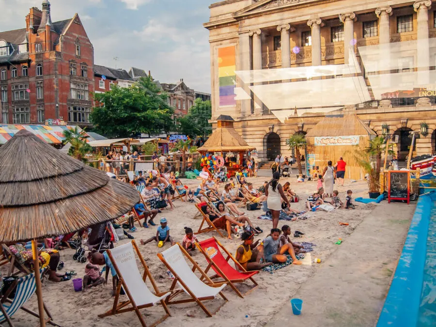Busy urban beach with visitors relaxing on deck chairs and playing in sand, surrounded by historic buildings and a paddling pool.