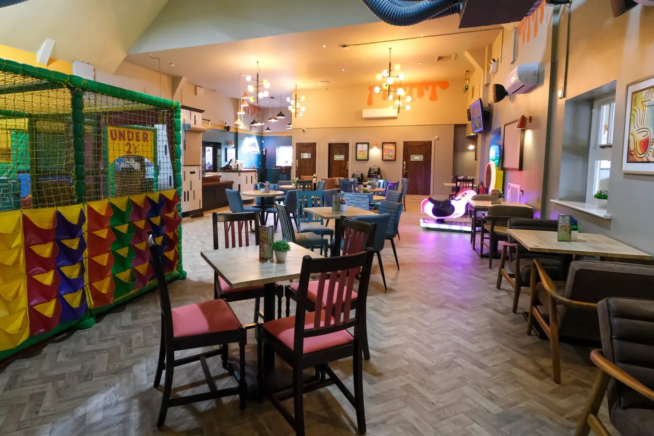 Spacious indoor soft play venue with dining tables, chairs, a colourful ball pit structure on the left, and a ride-on toy in the background.