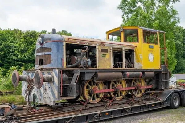 Vintage industrial steam locomotive with blue and yellow cab on a railway transport trailer, surrounded by greenery.