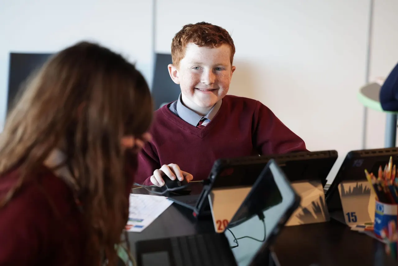 Boy and girl working together at computer stations with tablets in a classroom setting.