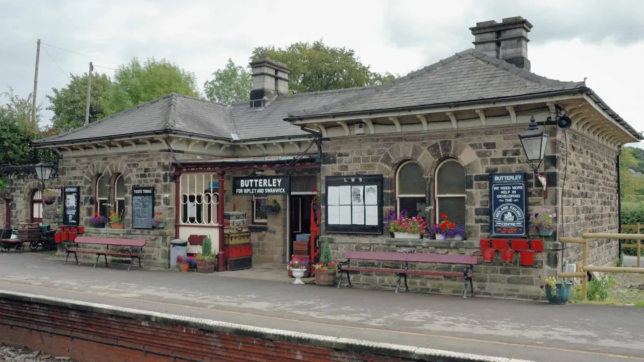 Stone railway station building with red benches, flowering planters, and period signage on the platform.