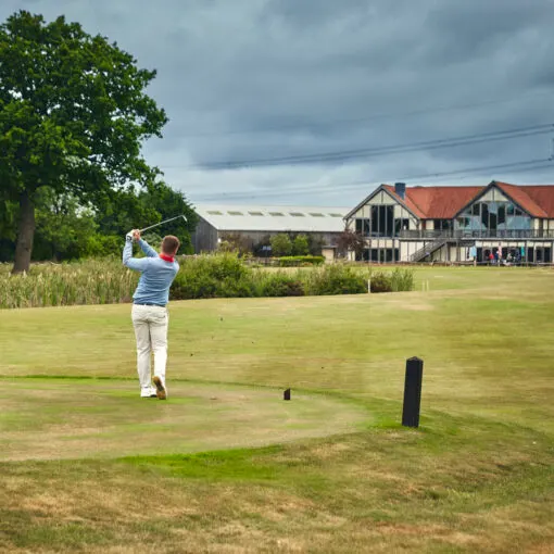 Golfer mid-swing on a fairway with Tudor-style clubhouse and grounds in the background.