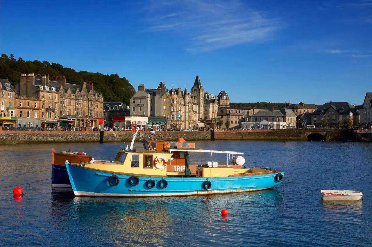 A colourful fishing boat moored in a Scottish coastal harbour with traditional stone buildings and waterfront shops lining the shore.