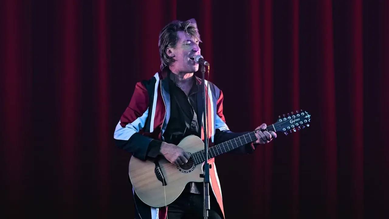 Male musician performing on stage with acoustic guitar at microphone against red curtain backdrop.