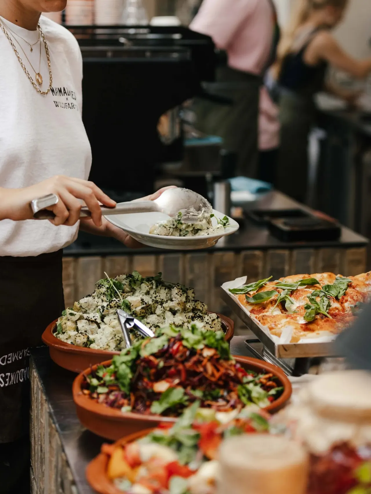 Person serving food at a buffet with pizza, salads and other dishes displayed on the counter.