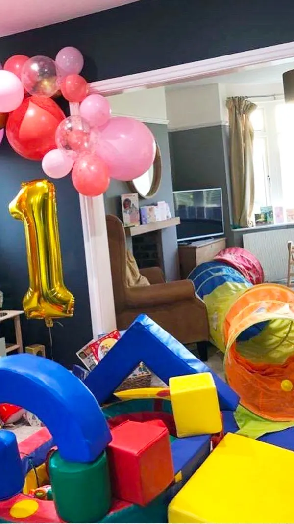 A children's play area decorated with pink and red balloons, a gold number one foil balloon, and colourful soft play equipment including tunnels, foam blocks and cushions.
