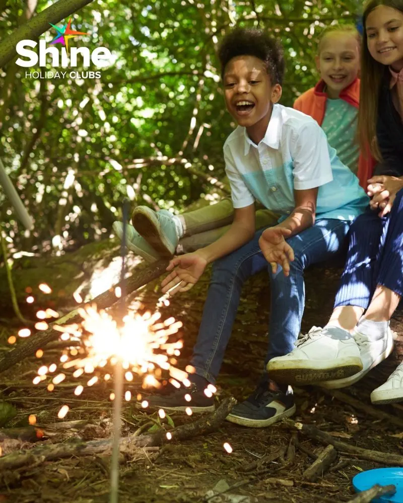 Children sitting on a tree branch watching a sparkler light up in the forest.