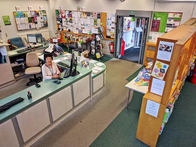 Staff member sits at reception desk in a busy library with notice boards, books and information displays visible.