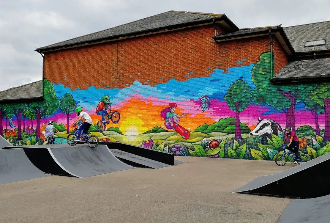 Colourful mural on brick building above BMX skate ramps showing children on bikes and scooters in a vibrant outdoor landscape scene.