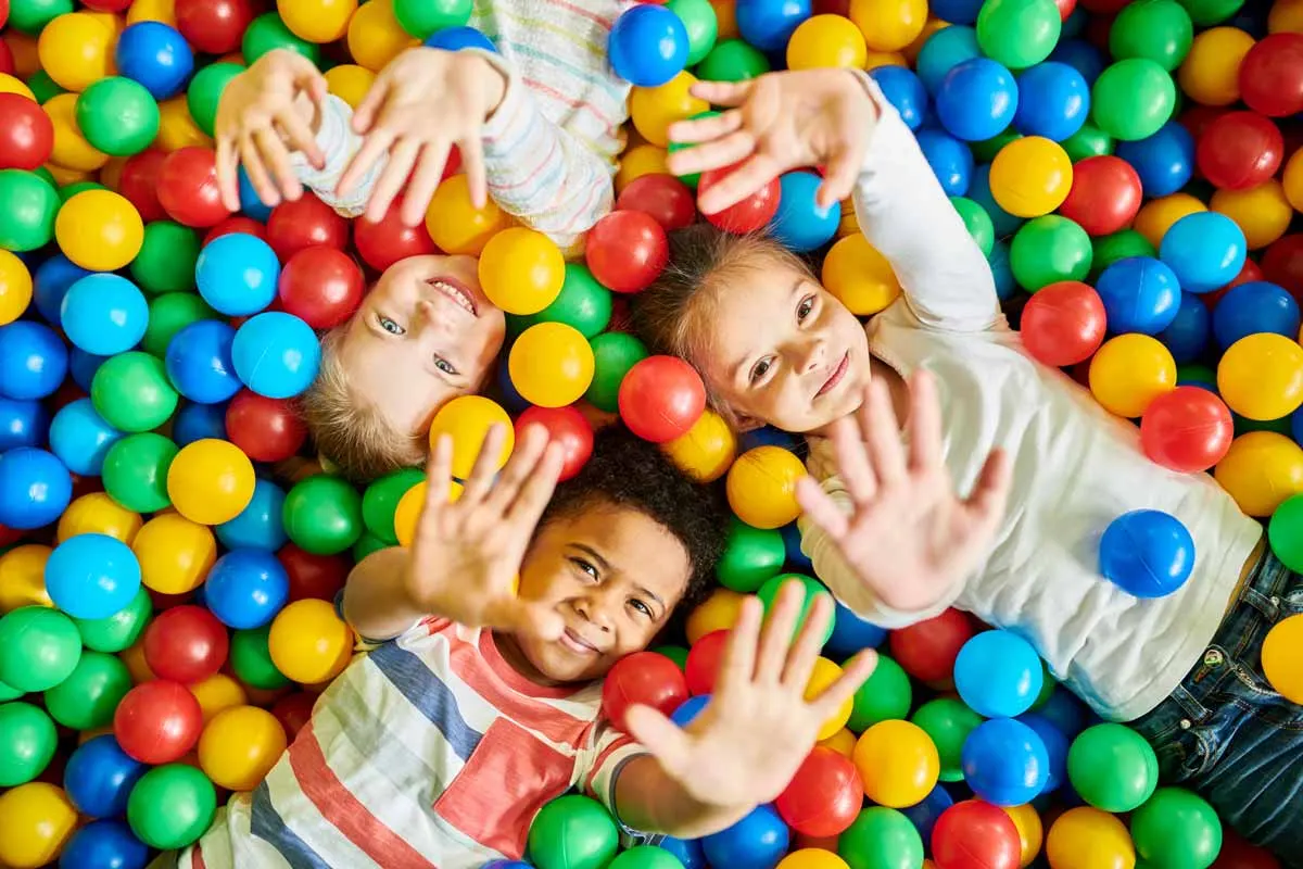 Three children lying in a ball pit filled with colourful plastic balls, smiling at the camera.
