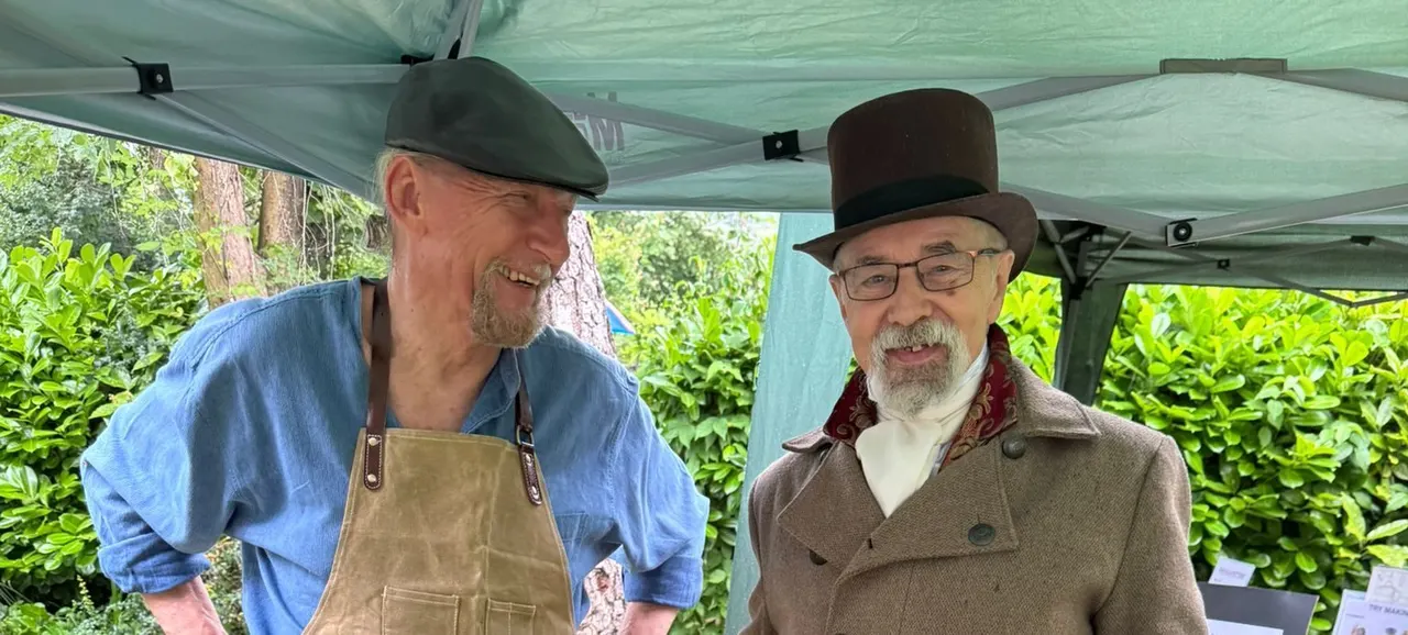 Two men in period costume stand together under a green marquee, with lush green foliage visible behind them.