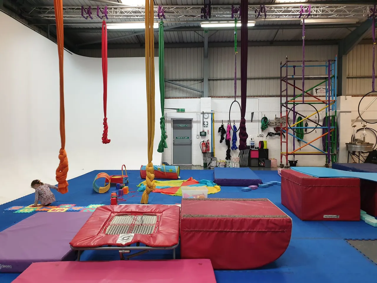 A young child plays on a blue padded floor surrounded by colourful climbing ropes, tunnels and soft play equipment in an indoor aerial arts studio.