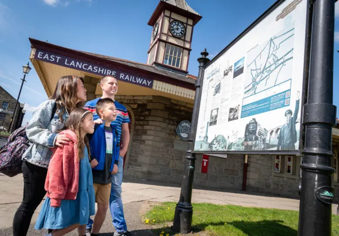 Two adults and two children viewing an information board at a heritage railway station with a clock tower.