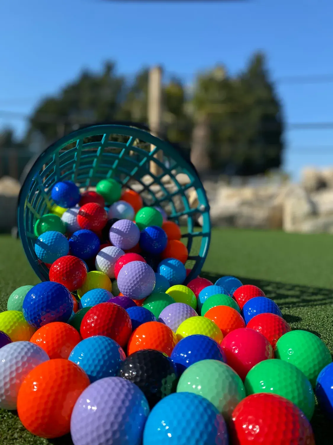 Colourful golf balls spilling from a tipped basket onto artificial turf, with ruins visible in the blurred background.