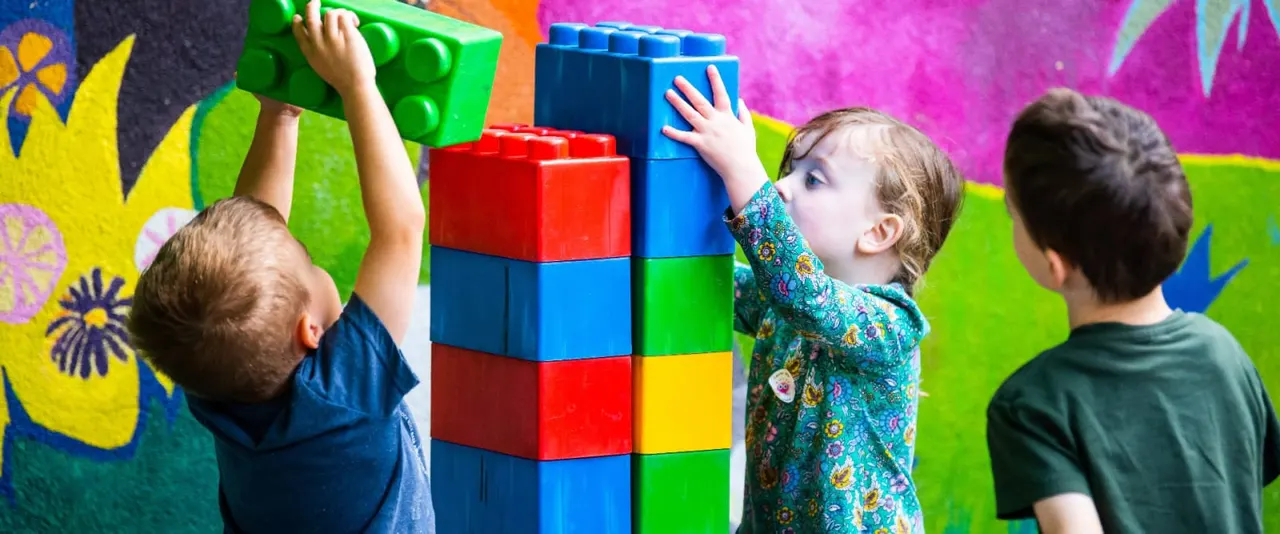 Three young children playing with oversized colourful building blocks against a vibrant painted wall.
