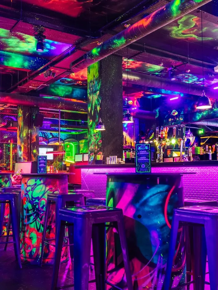 Neon-lit arcade bar interior with glowing pink, green and blue lighting, gaming machines, bar counter and seating areas decorated with vibrant graffiti-style artwork.