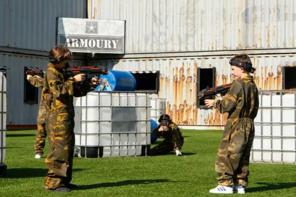 Three people in camouflage suits playing laser tag outdoors on grass near industrial buildings.