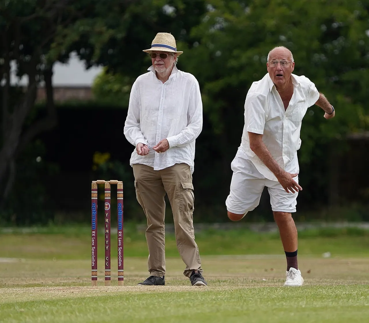 Two older men playing cricket on a grass pitch, one acting as bowler and one as batsman, with stumps and hedges visible behind.