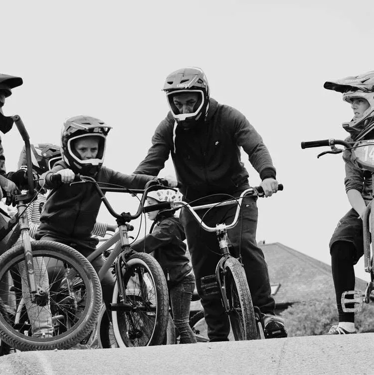 An adult and three children wearing protective helmets riding BMX bikes outdoors on a dirt track.