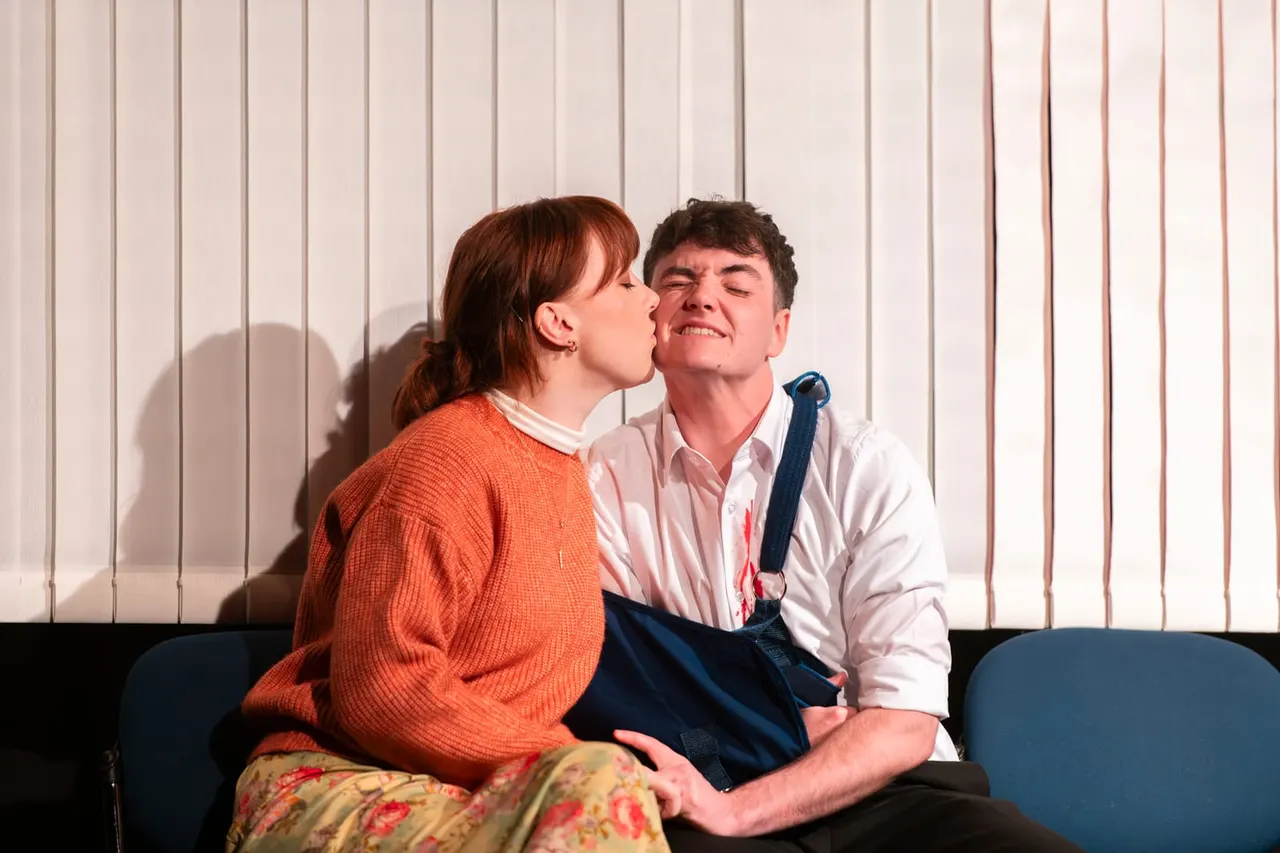 Two young performers on stage sitting on a blue seat, one kissing the other's cheek, against a white panelled backdrop.