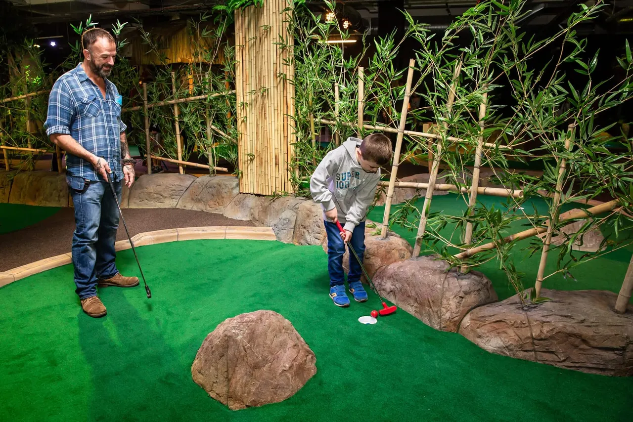 Adult and child playing mini golf on a green course with rock features and bamboo plants.