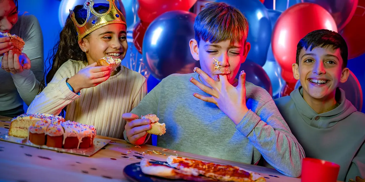 Three children eating cake at a colourful birthday party with balloons and blue and red lighting.