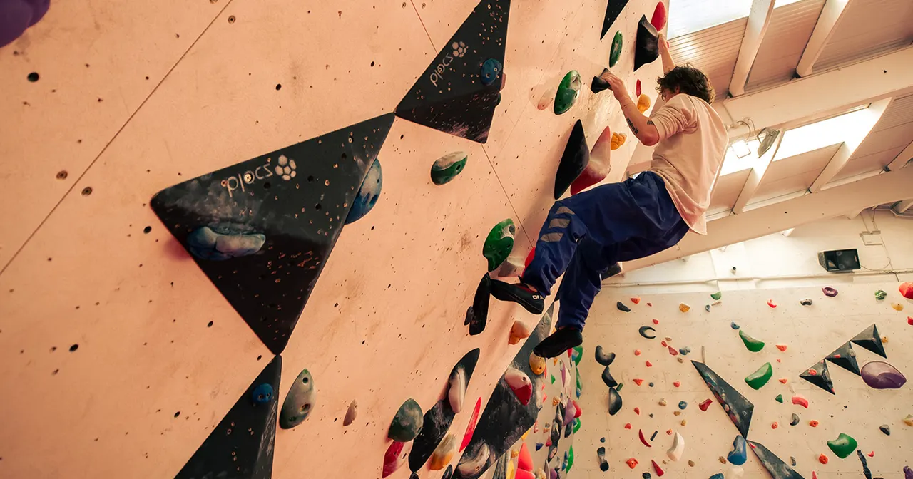 A person climbing an indoor climbing wall with multicoloured handholds beneath bright ceiling lights.