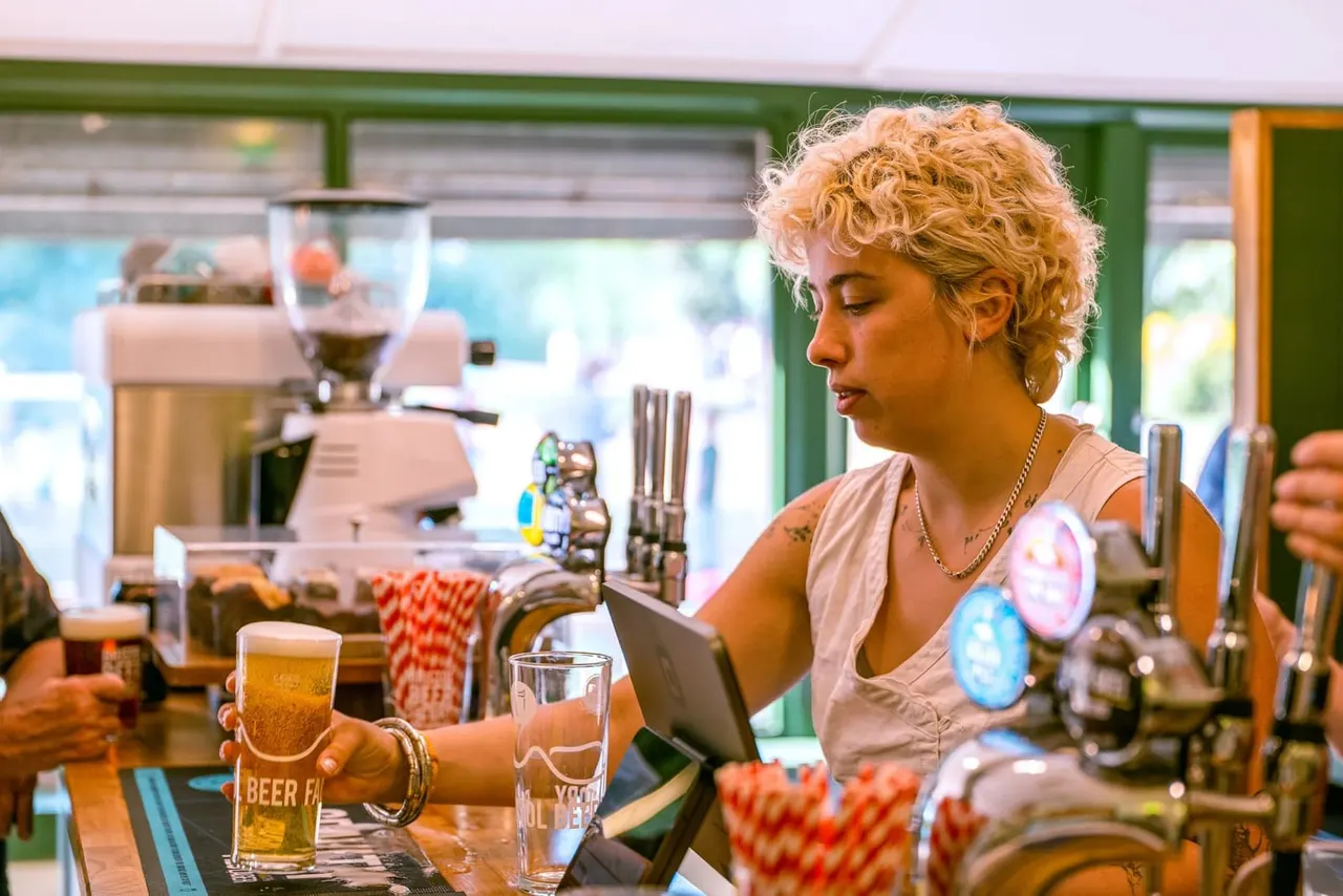 Barmaid serving a pint of beer at a busy bar counter with beer taps and glassware.