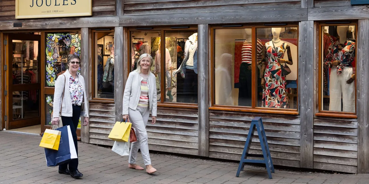 Two women carrying shopping bags walk past a fashion boutique with wooden frontage and dressed mannequins in the windows.