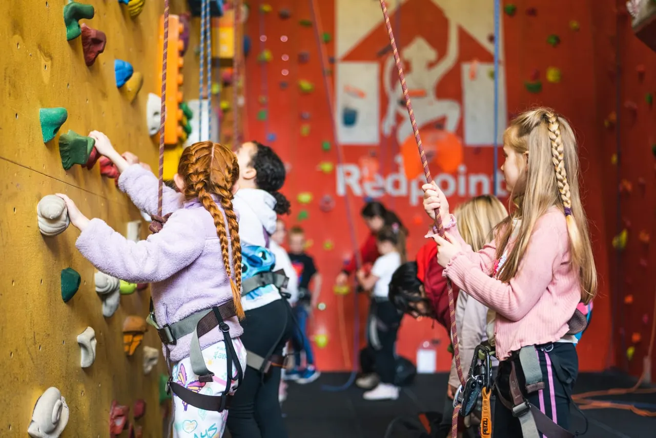 Children climbing coloured indoor climbing walls with safety harnesses whilst others watch in the background.