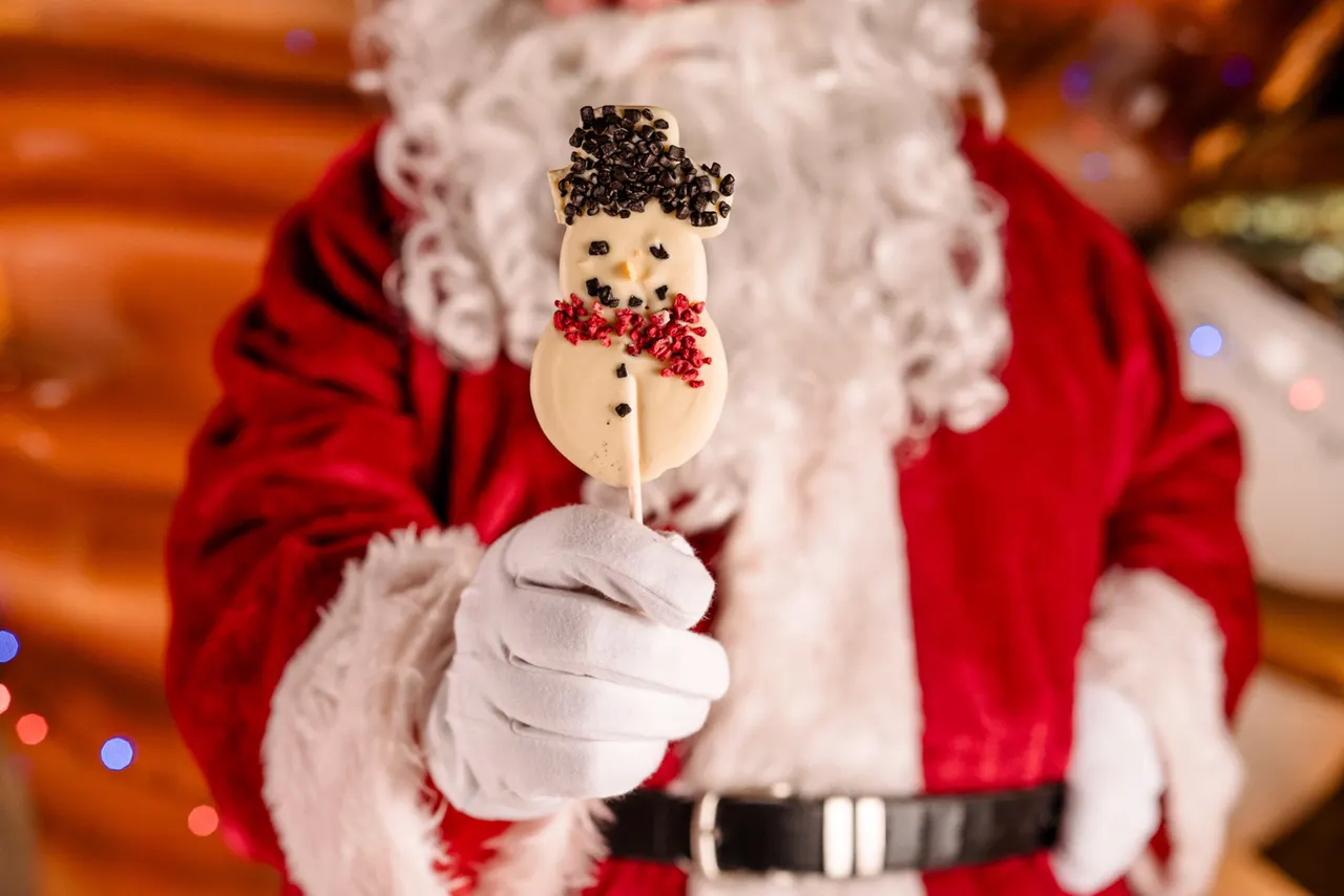 Person in Father Christmas costume holding a snowman-shaped chocolate lollipop with chocolate chips and red bow.