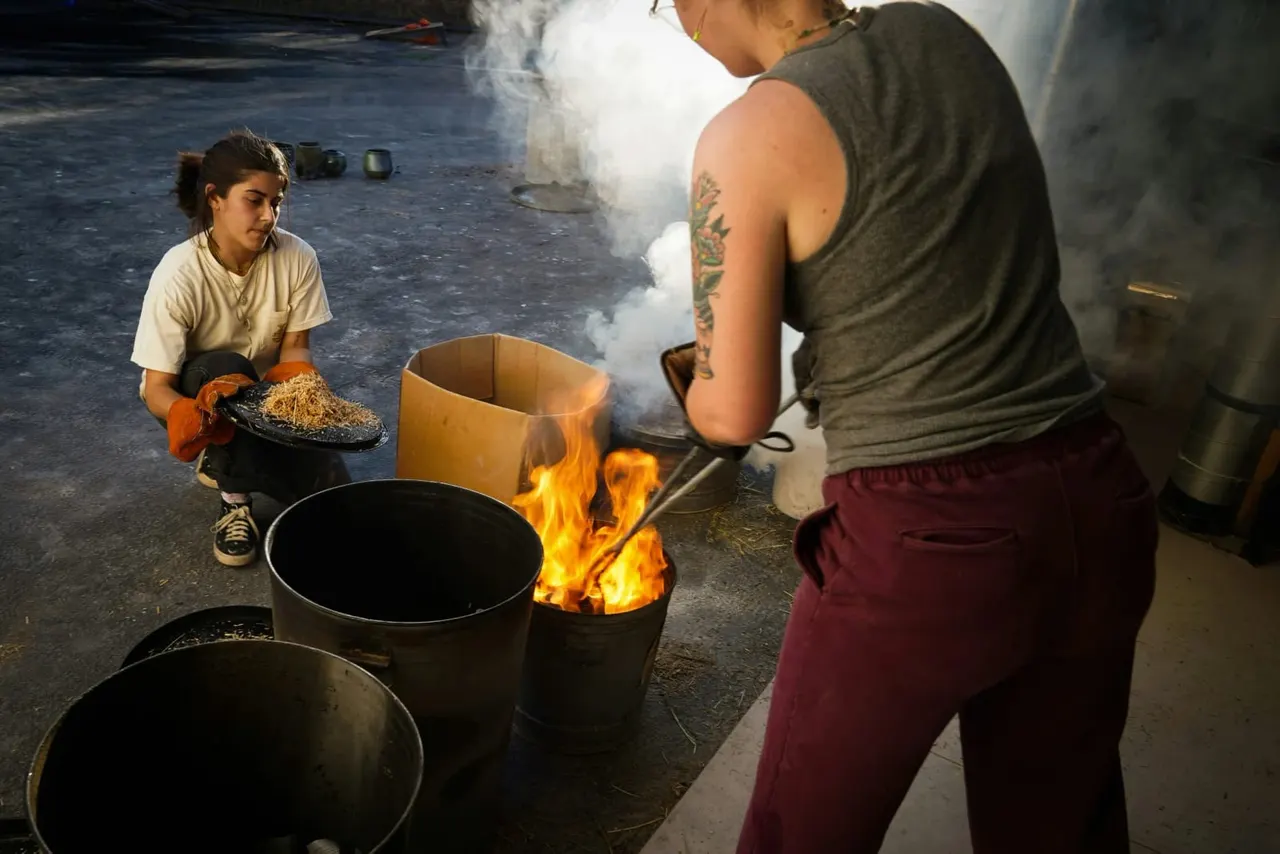 Two people cooking over an open fire in a pottery workshop, with flames and steam rising from metal pots.