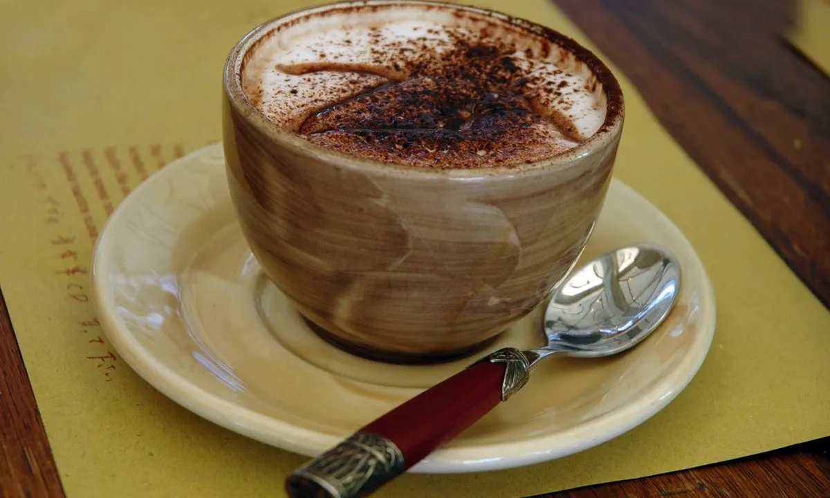 Cappuccino with cocoa powder dusting in a ceramic cup on a saucer with a spoon, on a yellow table.