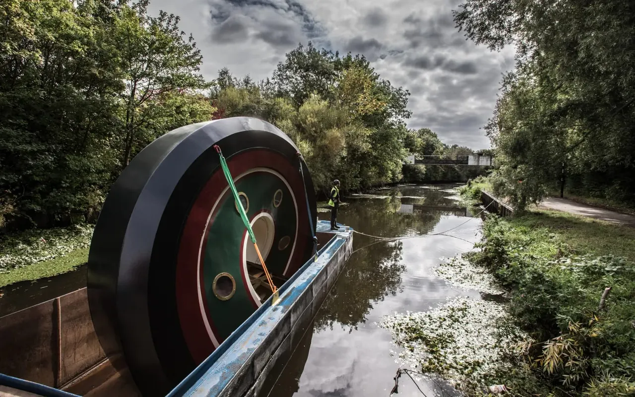 A person stands on a narrow boat featuring a large industrial water wheel, moored on a tree-lined canal.