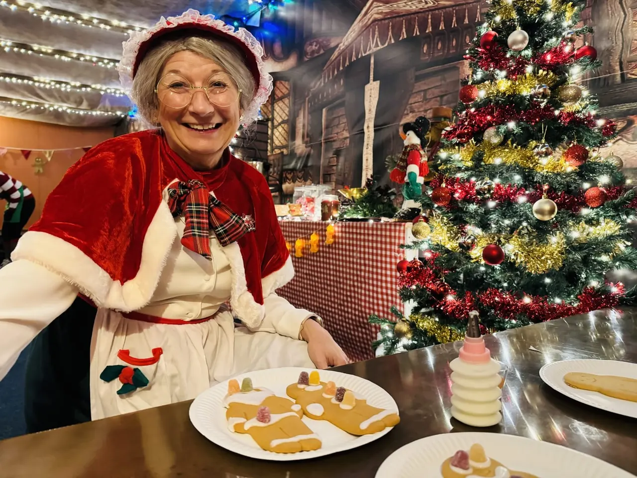 Woman in festive Christmas outfit posing at table with decorated gingerbread biscuits and Christmas tree backdrop.