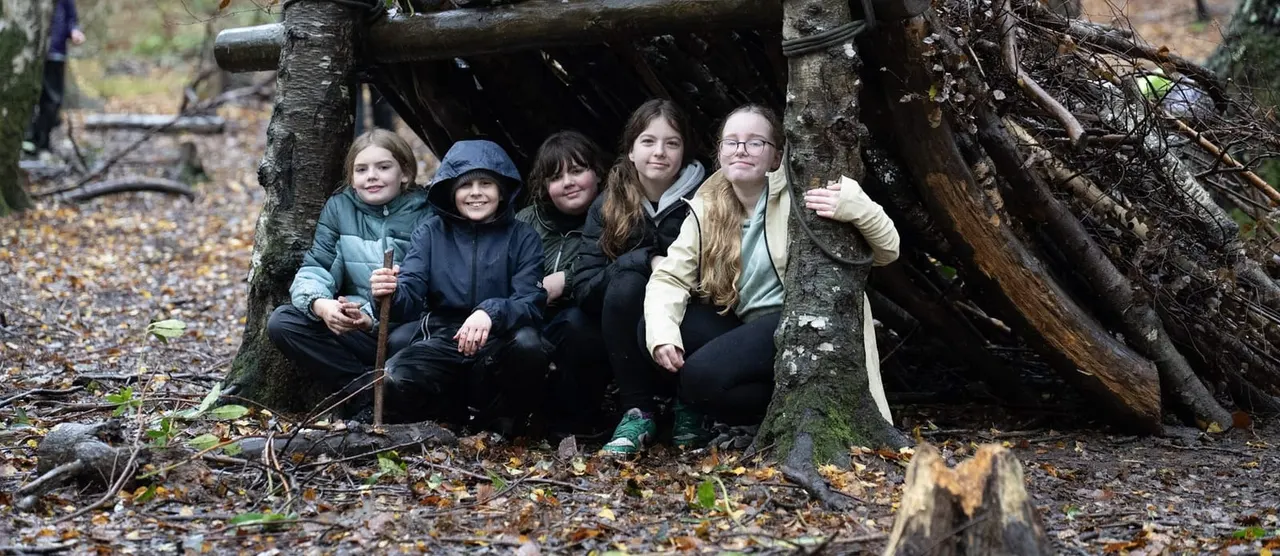 Five young people smiling inside a rustic wooden shelter made from logs and branches in a woodland setting.