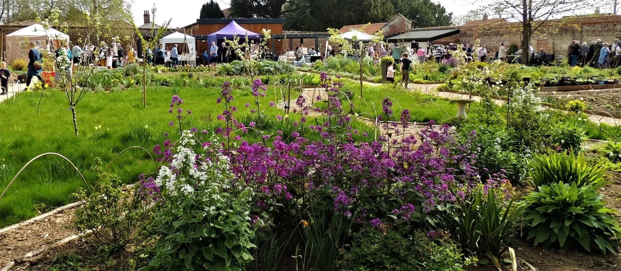 Visitors stroll through a garden venue with flowering beds including purple blooms, whilst marquees and refreshment facilities operate in the background.