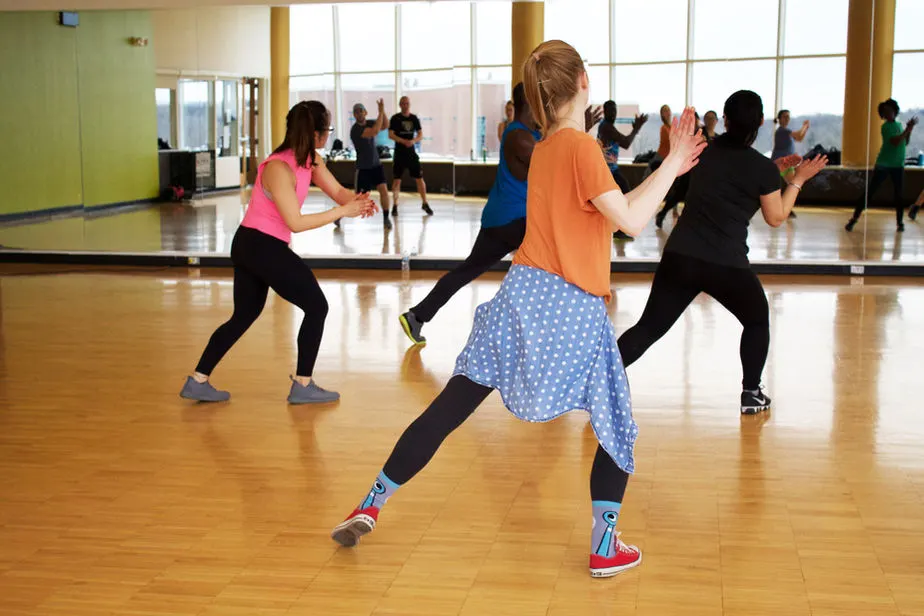 Group fitness class with participants doing aerobic exercise in a studio with mirrored walls and wooden flooring.