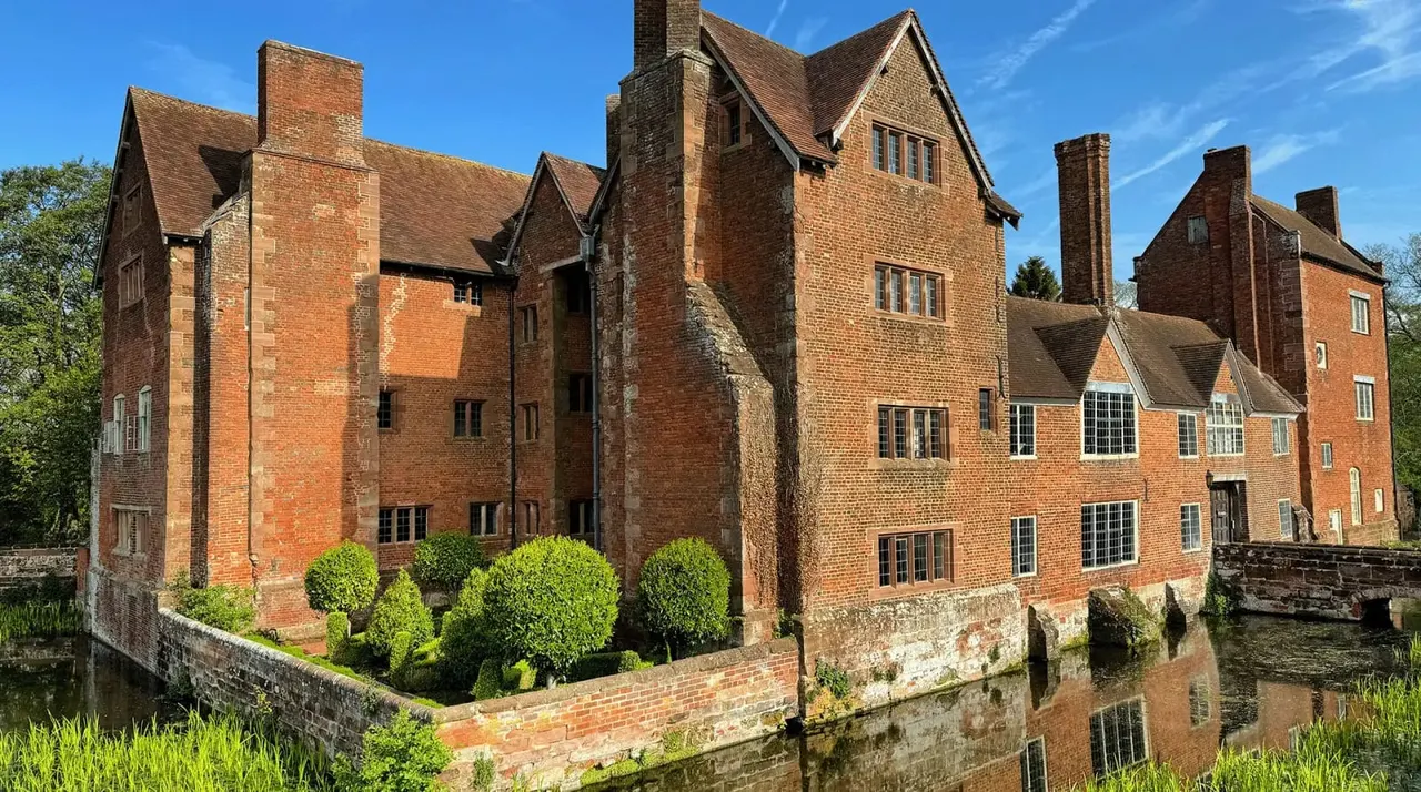 A historic brick manor house with multiple chimneys and gabled roofs surrounded by manicured gardens and a moat under clear blue sky.