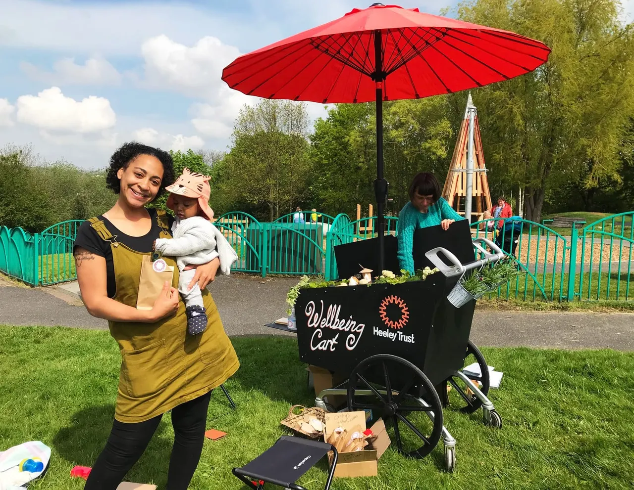 A woman holding a toddler stands next to a Wellbeing Cart under a red umbrella at a play park on a sunny day.