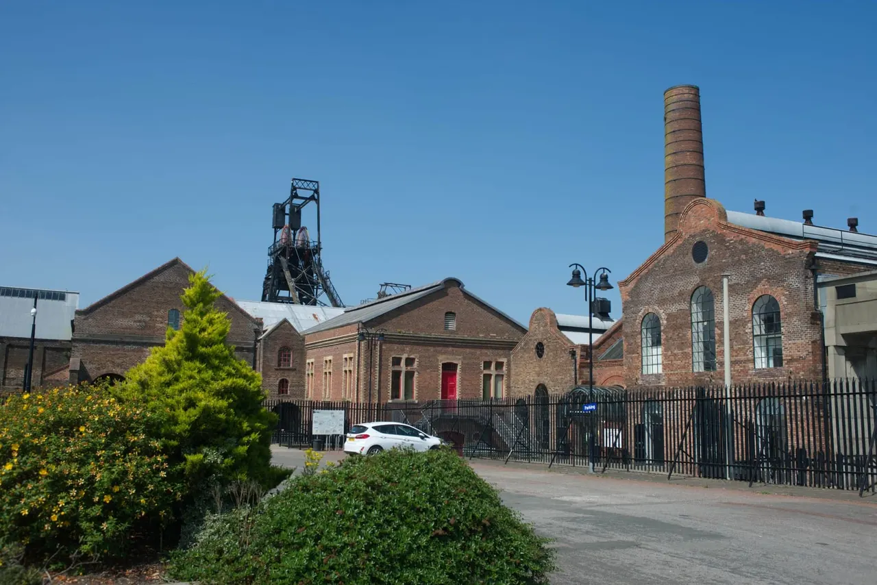 Historic red brick mining buildings with tall chimney and pit head gear under clear blue sky.