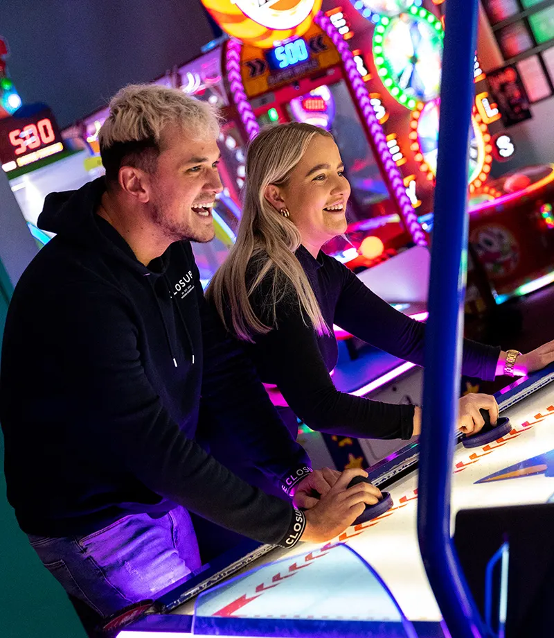 A man and woman play arcade games surrounded by brightly lit gaming machines in a leisure venue.