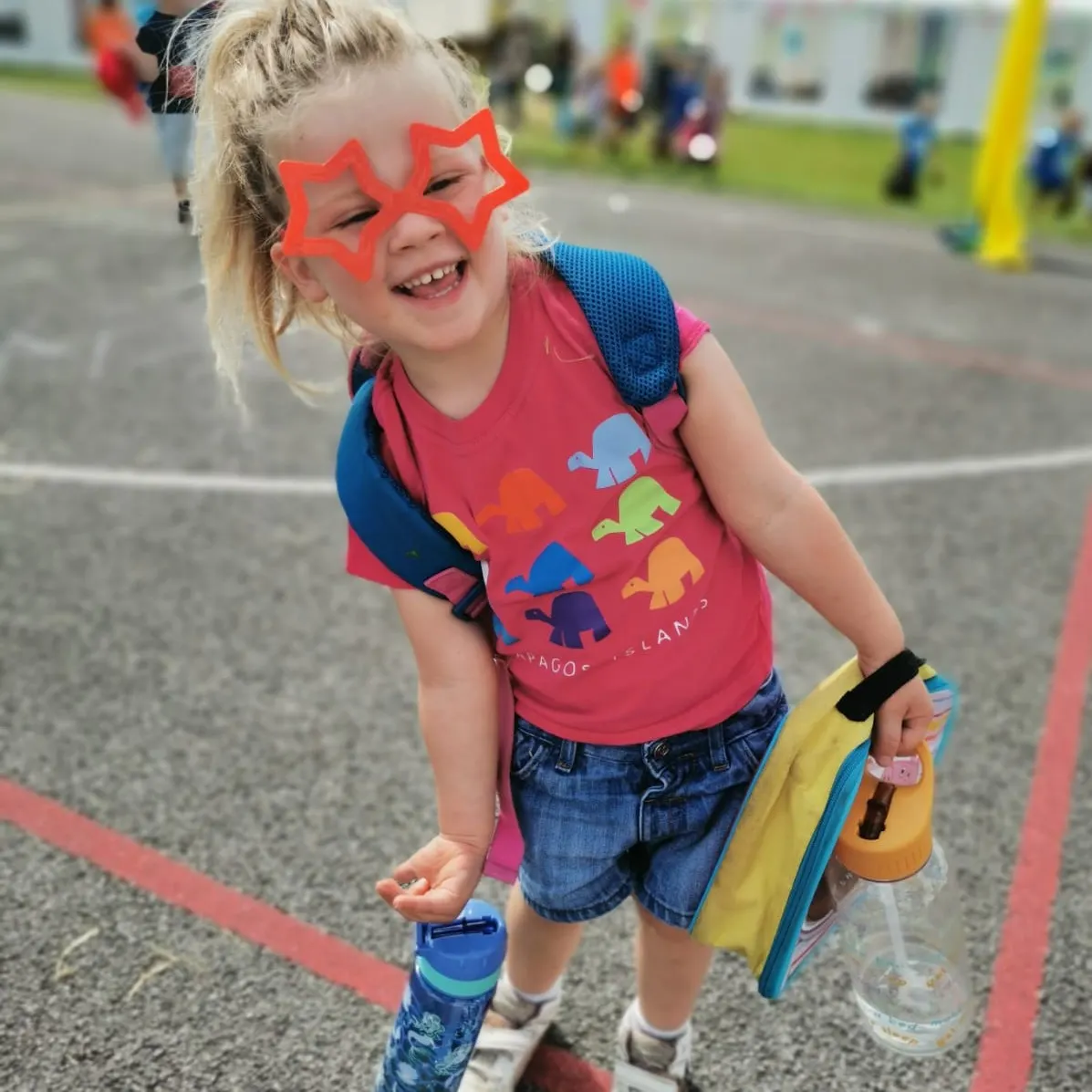 A young child wearing large red star-shaped glasses and a colourful dinosaur T-shirt smiles at a leisure park, holding a yellow bucket and activity items.