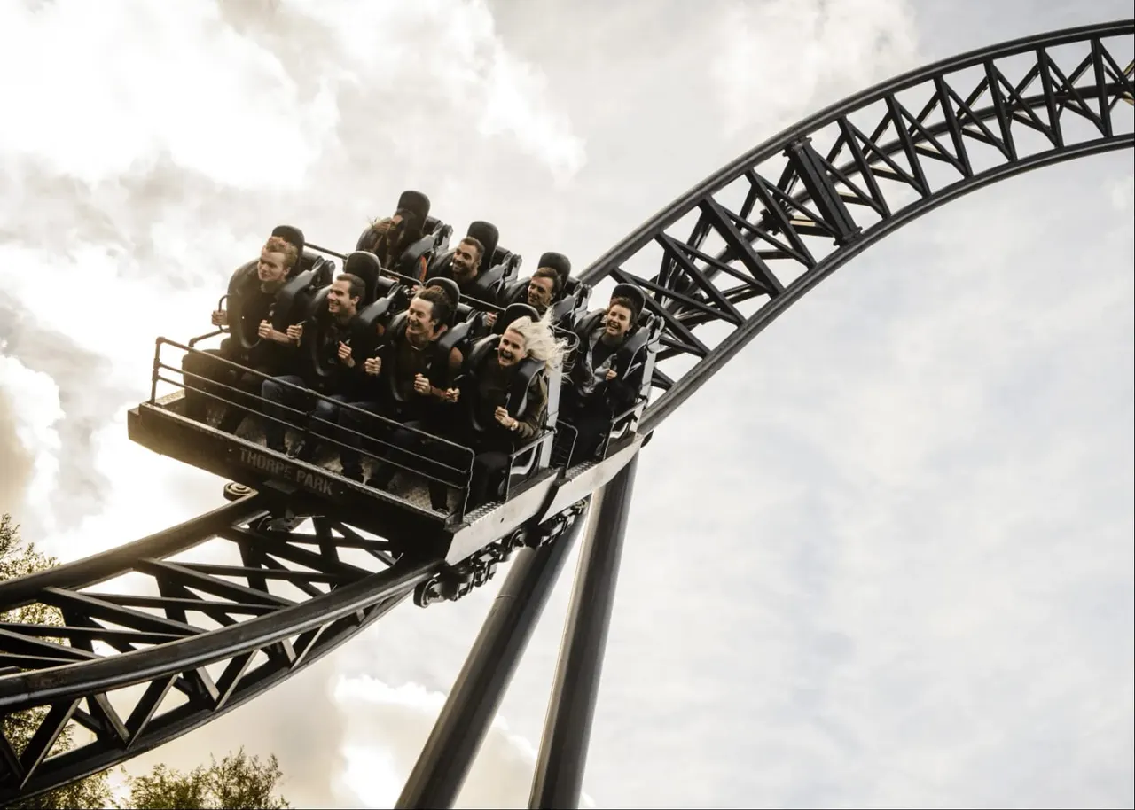 A group of people ride a roller coaster car ascending a steep metal track against a cloudy sky.