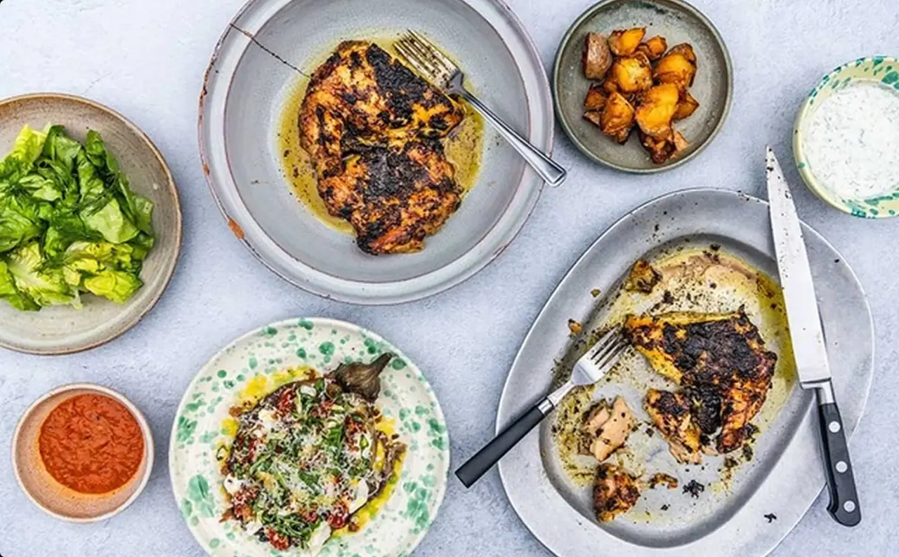 Two grilled fish fillets served with roasted potatoes, salad, white sauce, and red pepper dip on various plates.