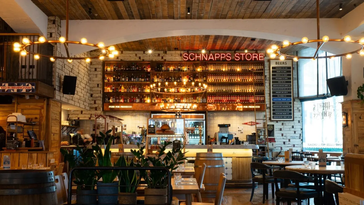 Interior of a German-style beer hall with illuminated bottle display, wooden beams, dining tables, and a service counter.