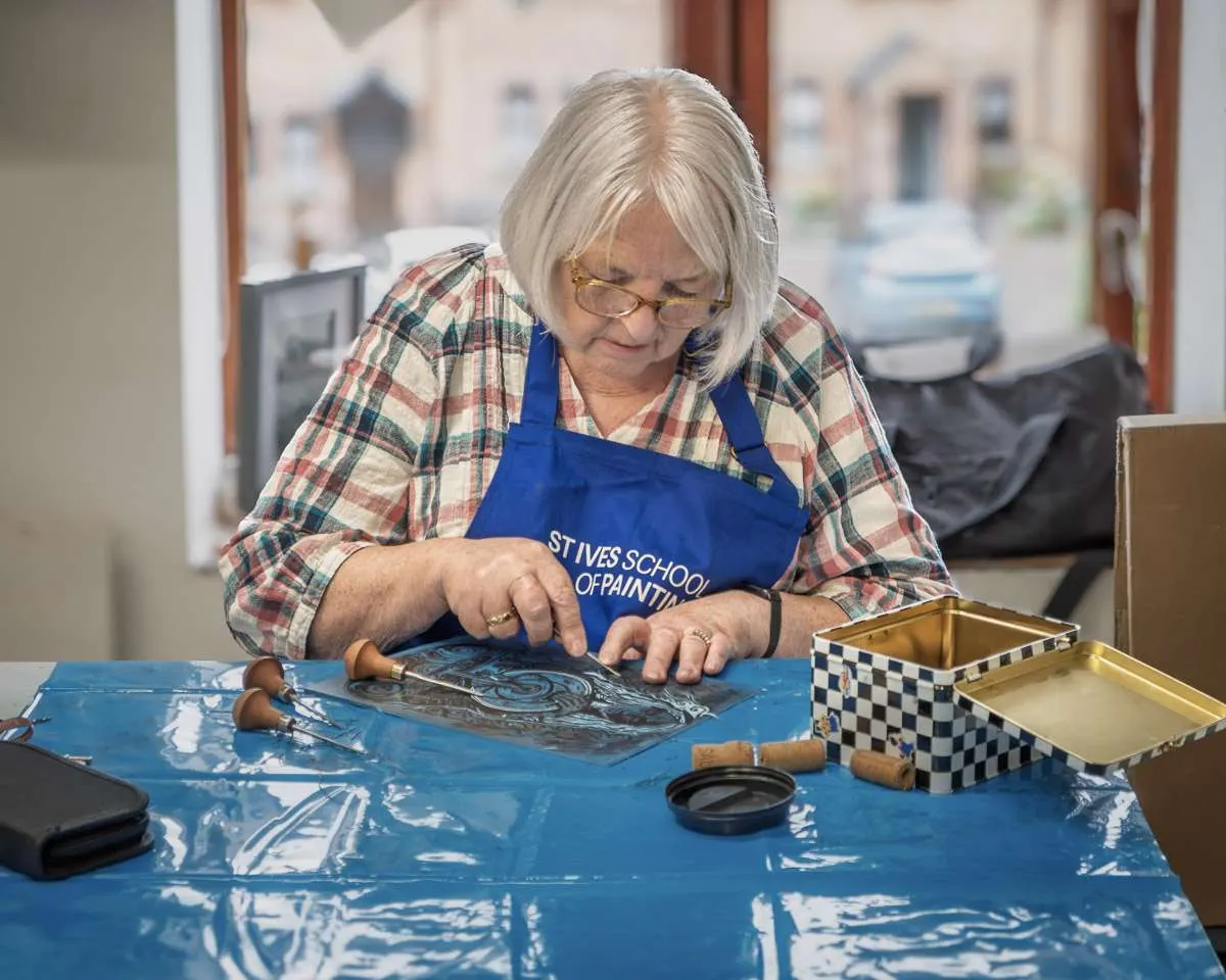 An older woman wearing glasses and a blue apron works on a blue printing plate at a table during a painting class.