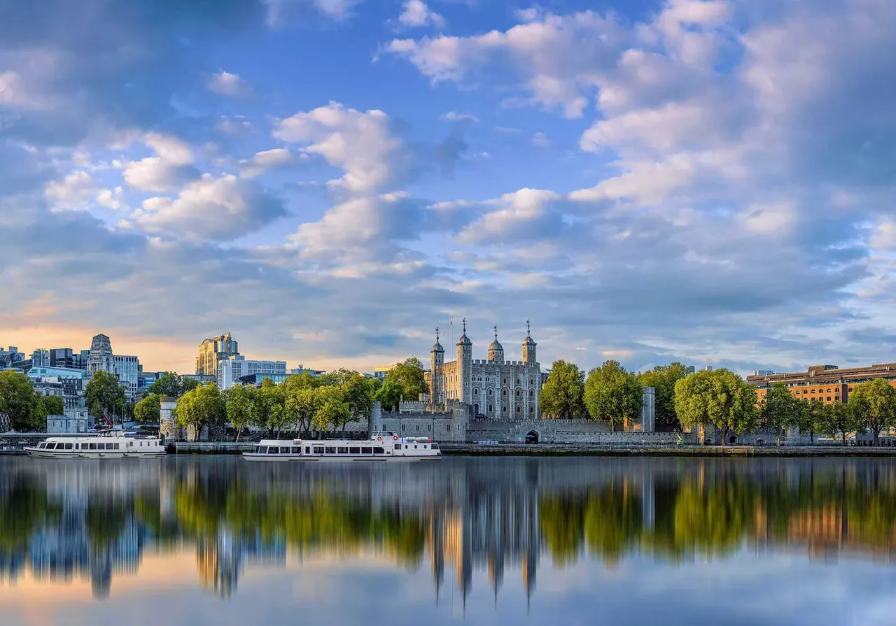 Tower of London and surrounding cityscape reflected in calm Thames river waters under blue sky with clouds.