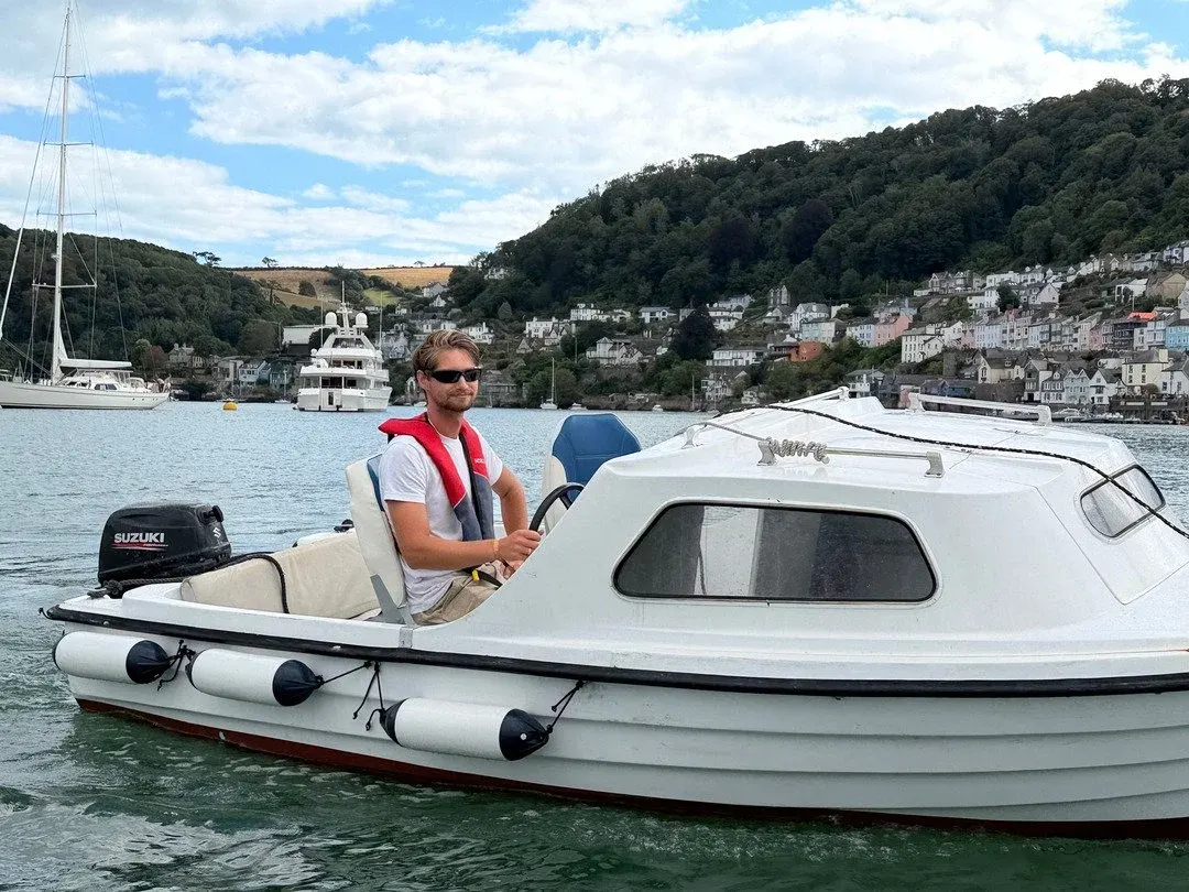 Man in red life jacket sits in white motorboat on river with wooded hillside and coastal town buildings behind.